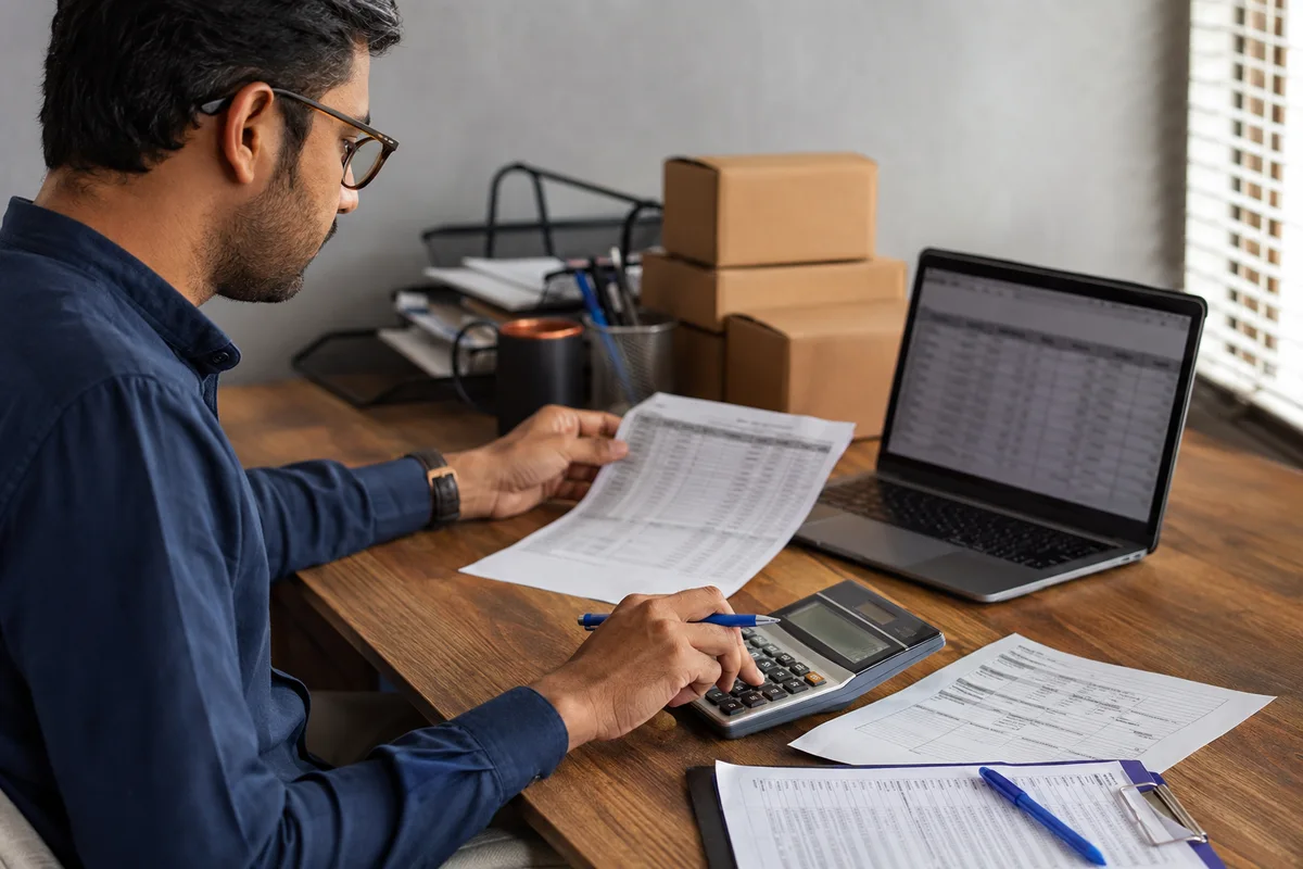 Procurement analyst checking quotation documents beside a calculator, laptop and sample cartons